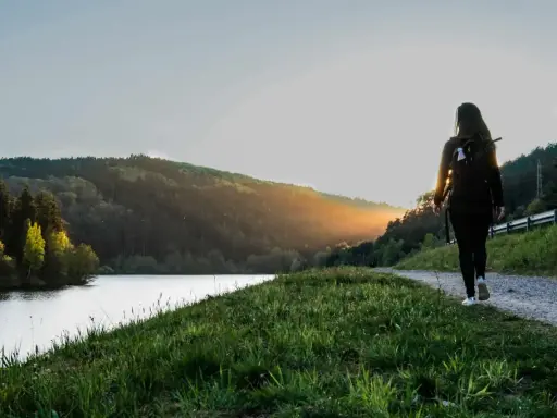 Frau mit Rucksack wandert auf einem Weg am See im Odenwald. Sonnenuntergang beleuchtet die Hügel.