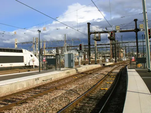 Ein weißer Hochgeschwindigkeitszug steht am Bahnsteig. Die Gleise führen in die Ferne, vorbei an Signalen und Oberleitungen. Vielleicht ist dies der **Zug nach Barcelona**?
