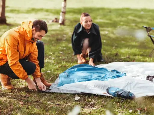 Ein Mann in einem orangen Anorak und eine Frau im Hintergrund bauen ein Zelt auf. Sie bereiten sich auf das **Zelt reinigen** nach dem Camping vor.
