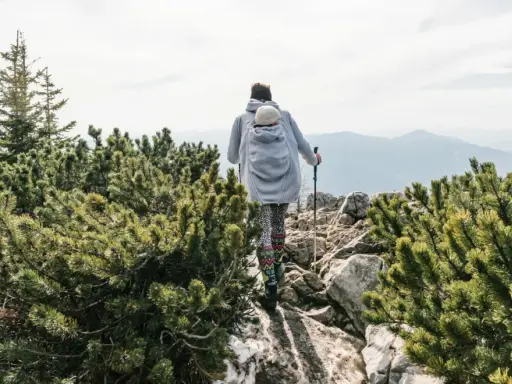 Frau wandert mit Baby im Tragetuch auf einem felsigen Bergpfad, umgeben von grünen Kiefern.