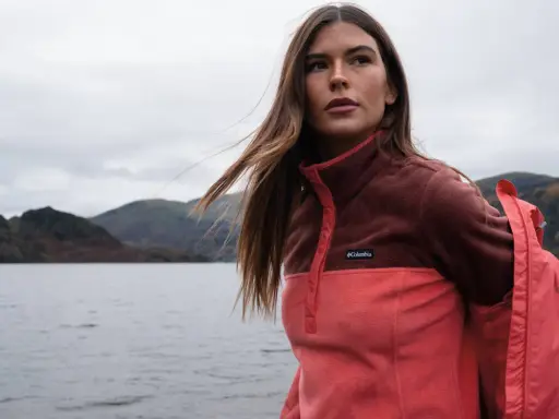 Frau mit langen Haaren im Zwiebel-Look, trägt eine zweifarbige Fleecejacke von Columbia am Seeufer.