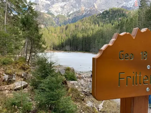 Holzschild "Geo 18 Fritter" vor gefrorenem See und Bergkulisse. Perfekt für einen Sommerurlaub in Bayern.