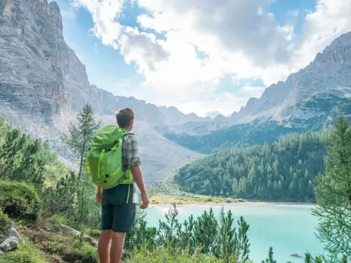 Wanderer mit Rucksack blickt auf türkisfarbenen Bergsee. Die Alpenlandschaft erinnert an die Grenze zwischen Schweiz und Italien.