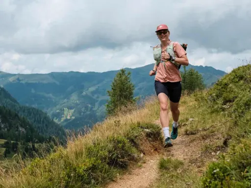 Lächelnde Frau beim Trailrunning Training auf einem Bergpfad, mit Blick auf grüne Täler und bewölkten Himmel.