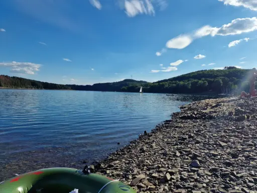 Sonniger Tag am See in NRW. Ein Segelboot gleitet über das Wasser, während am steinigen Ufer Menschen entspannen.