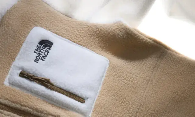 Ein beige-weißes Fleece-Oberteil von The North Face mit Reißverschlusstasche. Fleece ist ein weiches, warmes Material.