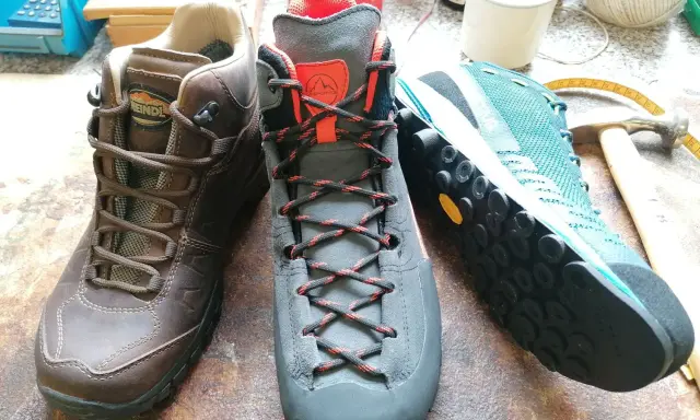 Drei Wanderschuhe von verschiedenen Herstellern: braun, grau und türkis. Werkzeuge und Garn liegen daneben.