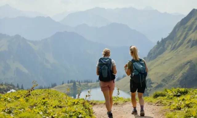 Zwei Wanderinnen genießen die Aussicht auf den **rundwanderwege Oberstdorf**. Ein Bergsee liegt im Tal, umgeben von sanften Hügeln und majestätischen Gipfeln.