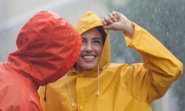 Zwei lachende Personen im Regen, eine in Gelb, die andere in Rot. Die gelbe Regenjacke ist im Fokus.