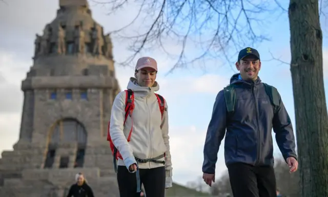 Ein Paar genießt eine Wanderung, passend mit wetterfester Wanderkleidung und Rucksäcken ausgestattet, vor einem imposanten Denkmal.