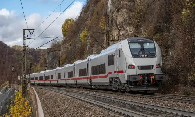 Ein weißer ICE-Zug mit roten Streifen fährt auf Gleisen an einer Felswand entlang. Herbstlaub färbt die Szenerie.