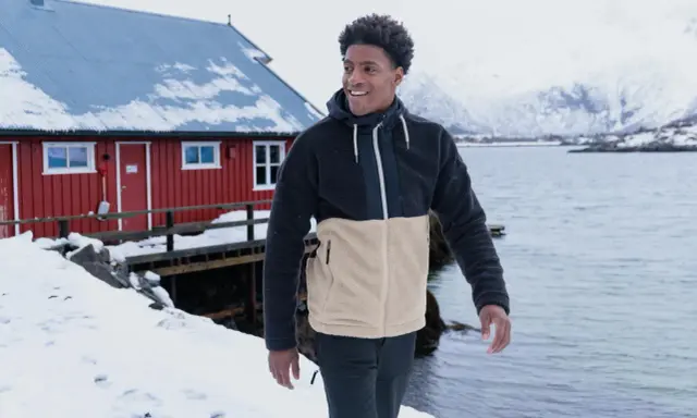 Mann in warmer Fleecejacke, bereit für das Waschen, steht vor einem roten Haus am Meer.
