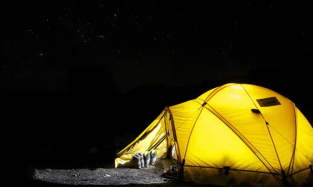 Gelbes Zelt leuchtet in der Nacht unter Sternenhimmel. Stiefel stehen bereit. Alles, was man zum Zelten braucht, ist hier.