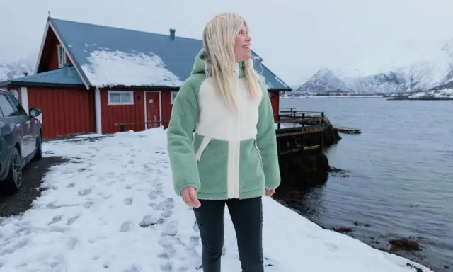 Frau in flauschiger Fleecejacke, bereit für das **fleece waschen**, steht vor rotem Haus am Meer.