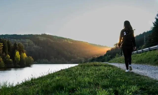 Frau mit Rucksack wandert auf einem Weg am See im Odenwald. Sonnenuntergang beleuchtet die Hügel.