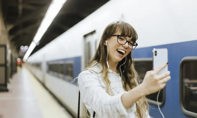 Junge Frau mit Kopfhörern macht ein Selfie am Bahnhof. Sie freut sich, ihr nächstes Abenteuer mit dem **discover eu pass** zu beginnen.
