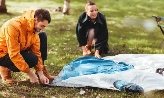 Ein Mann in einem orangen Anorak und eine Frau im Hintergrund bauen ein Zelt auf. Sie bereiten sich auf das **Zelt reinigen** nach dem Camping vor.