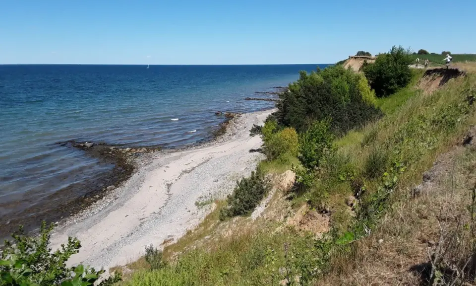 Ein sonniger Tag am Timmendorfer Strand. Die Ostsee glitzert, ein schmaler Kiesstrand lädt zum Spaziergang ein, während grüne Büsche die Steilküste säumen.