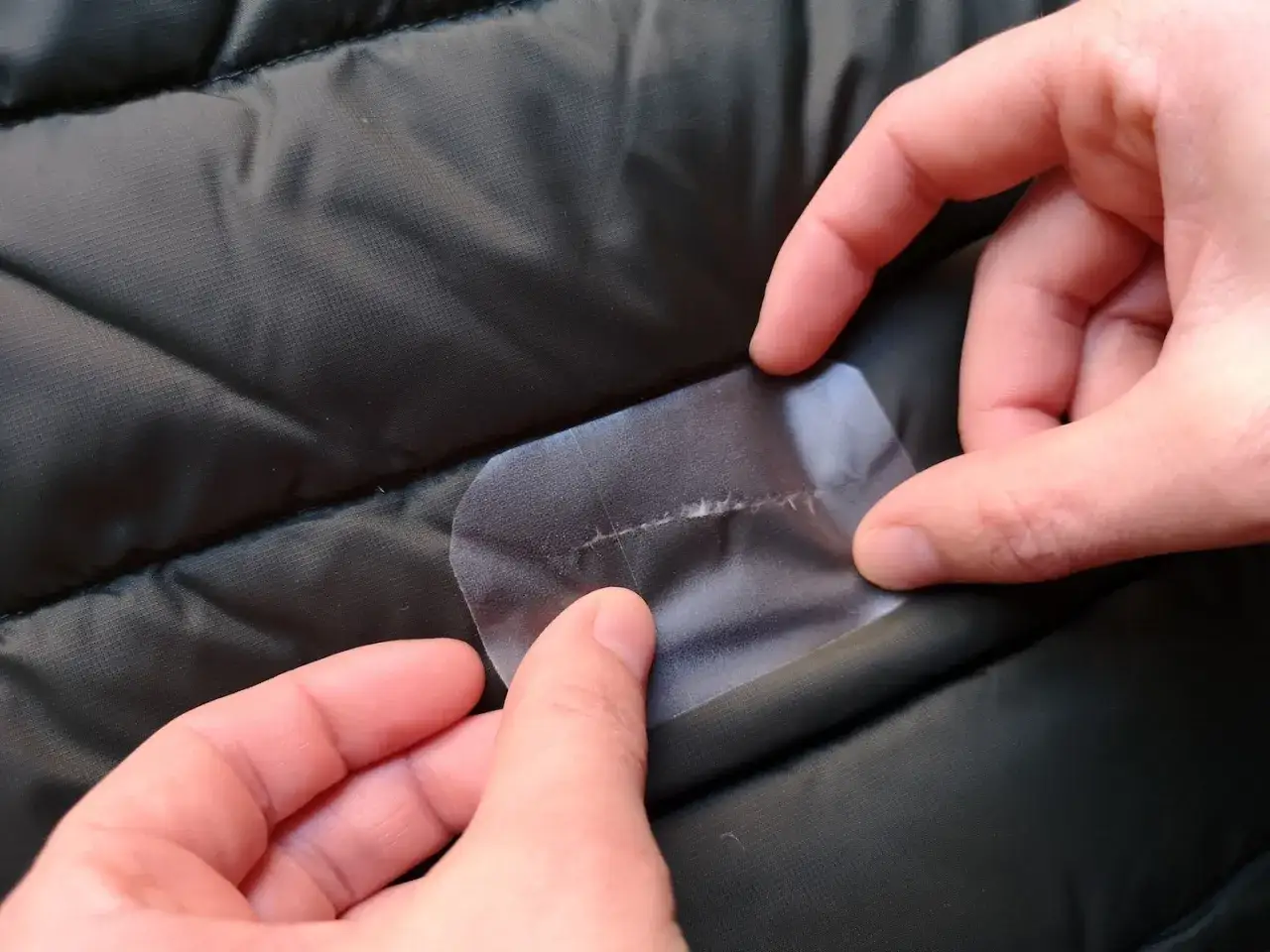 H&auml;nde reparieren eine Daunenjacke mit einem transparenten Flicken, der &uuml;ber einen Riss geklebt wird.
