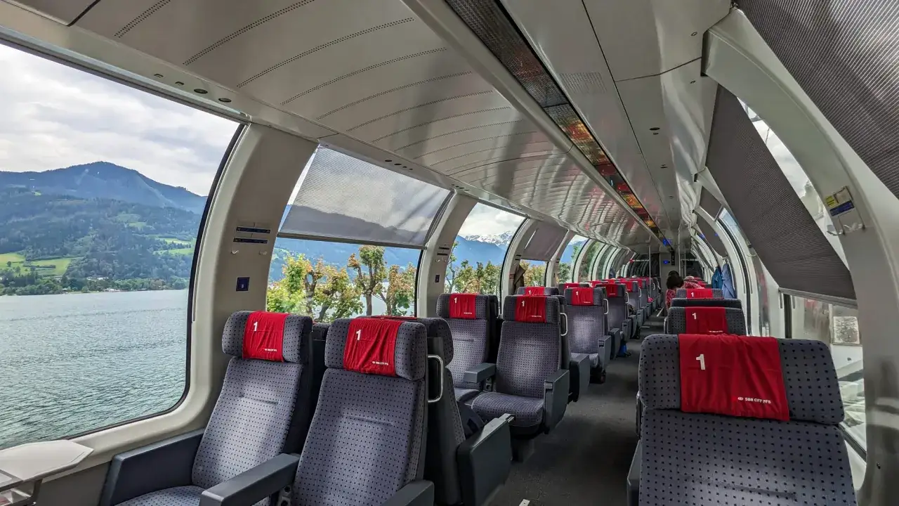 Innenansicht eines Panoramazugs in Deutschland mit Blick auf einen See und Berge. Die Sitze sind mit roten Kopfkissen mit der Nummer 1 versehen.