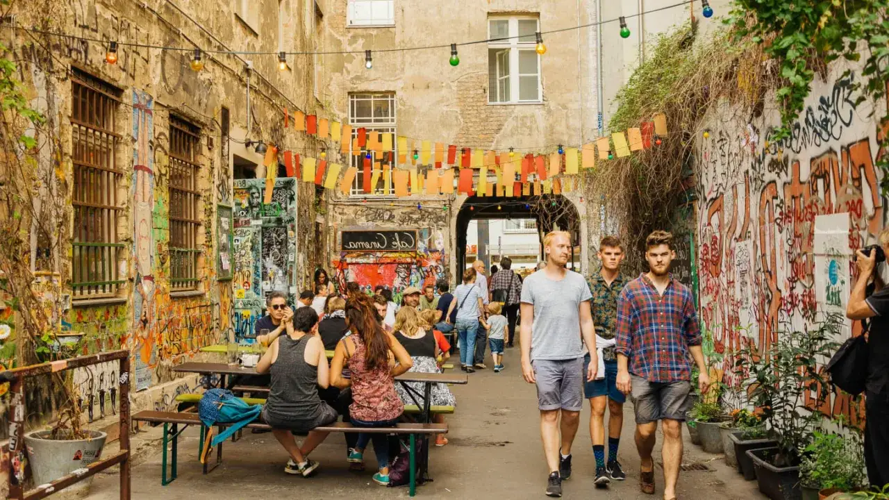 Bunte Lichterketten und Graffiti schmücken einen belebten Innenhof in Berlin Kreuzberg. Menschen sitzen an Tischen, genießen die Atmosphäre.