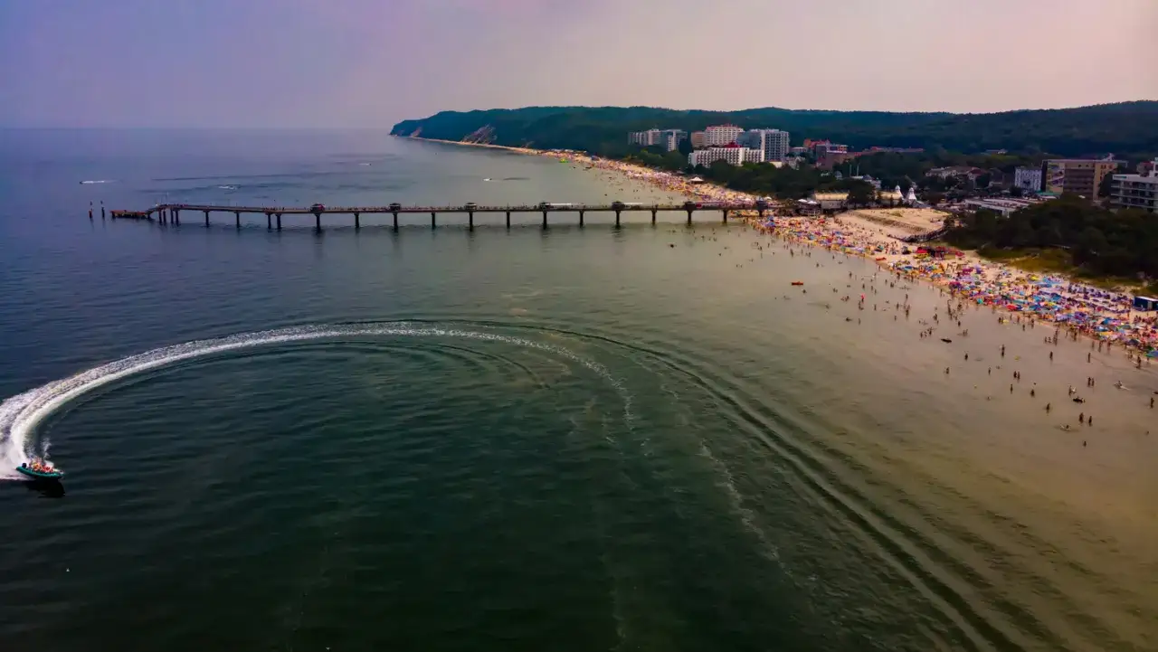 Ein belebter Strand in Misdroy, Polen, mit einer langen Seebr&uuml;cke und einem Schnellboot, das eine Spur im Wasser hinterl&auml;sst.