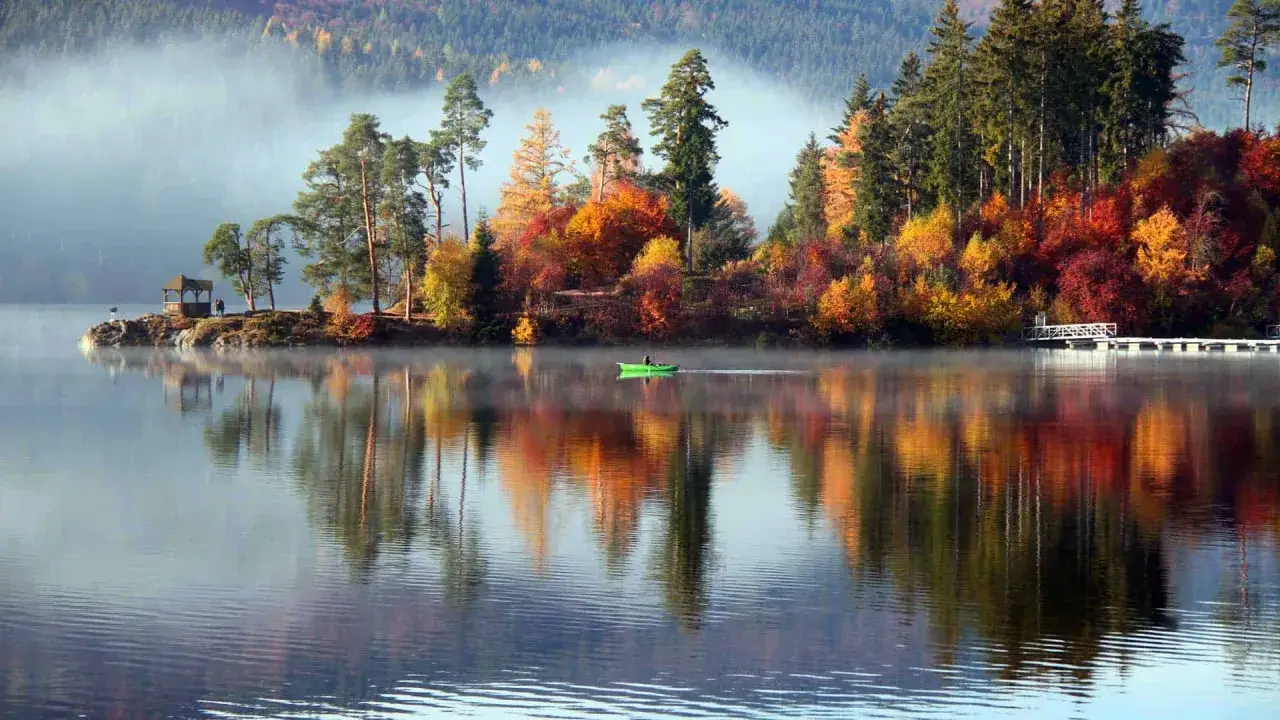 Ein ruhiger See im November, Nebel steigt auf. Ein kleines Boot gleitet &uuml;bers Wasser, bunte B&auml;ume spiegeln sich. Perfekt f&uuml;r einen Kurzurlab im November.
