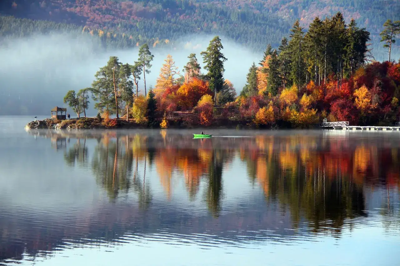Herbstlicher Geheimtipp Urlaub November Deutschland: Nebel &uuml;ber einem See mit bunten B&auml;umen und einem Ruderboot.