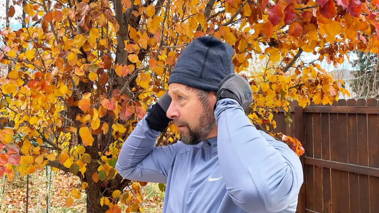 Mann mit Bart passt Mütze und Handschuhe an. Die bunte Herbstlaubkulisse deutet auf die richtige Kleidung zum Laufen im Winter hin.