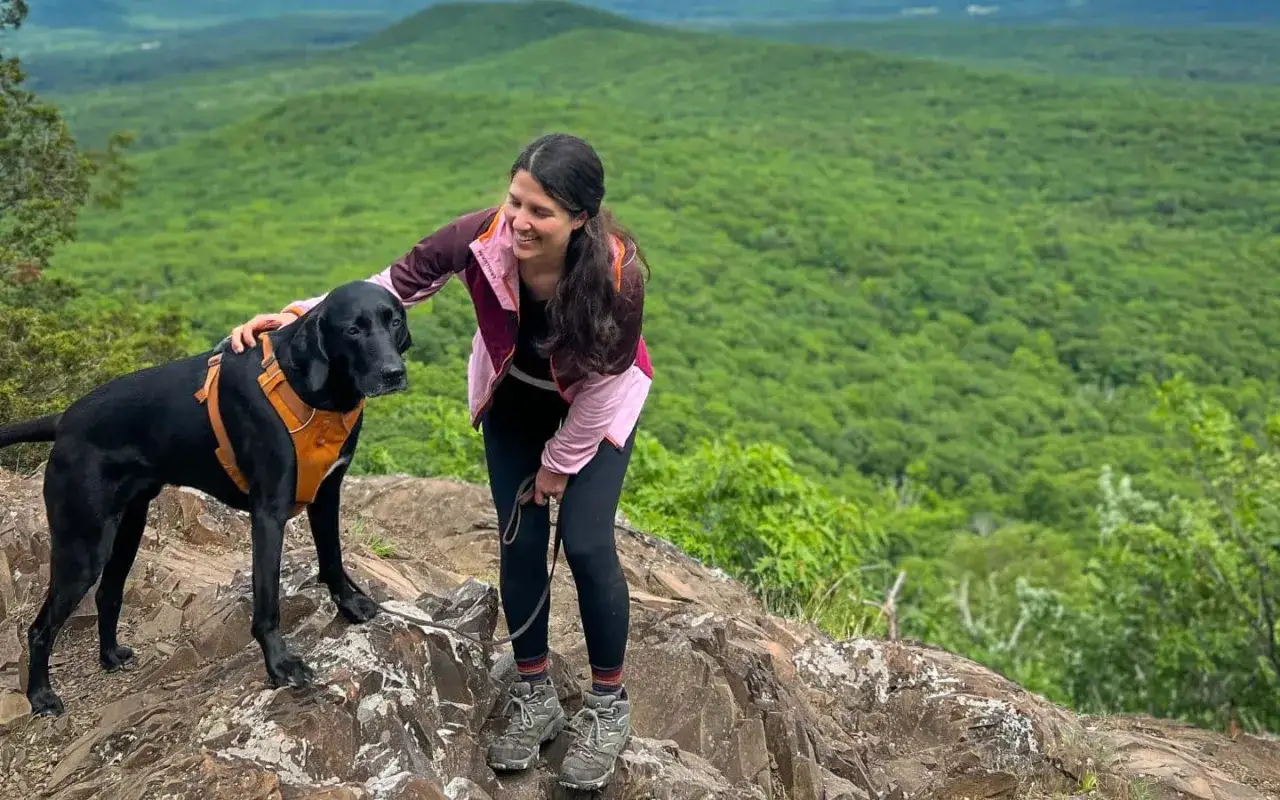 Frau streichelt ihren schwarzen Hund beim Wandern mit Hund auf einem felsigen Gipfel mit Blick auf einen grünen Wald.