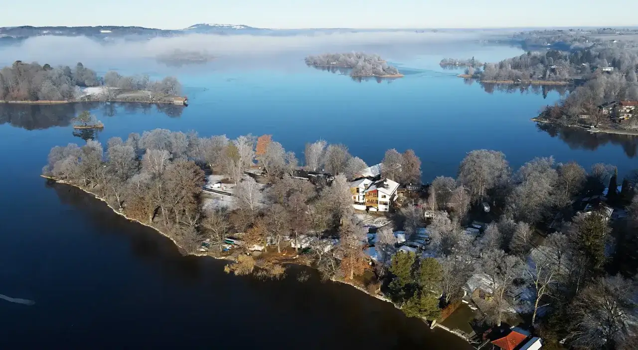 Winterliche Idylle am Staffelsee: Ein Haus mit roten Fensterl&auml;den auf einer schneebedeckten Halbinsel, umgeben von gefrorenen B&auml;umen und dem blauen See.