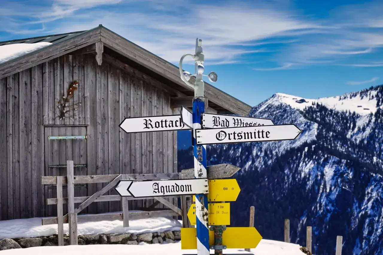 Wegweiser am Fockenstein Lenggries zeigt Richtungen nach Rottach, Bad Wiessee, Ortsmitte und Aquadom. Schneebedeckte Berge im Hintergrund.