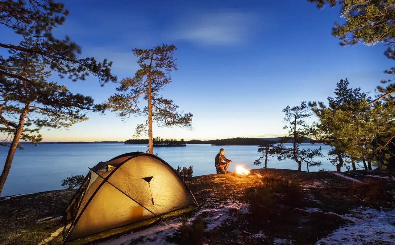 Ein Zelt und ein Lagerfeuer am Seeufer, ein Mann sitzt davor. Ein friedliches Bild von Wildcampen Finnland bei D&auml;mmerung.