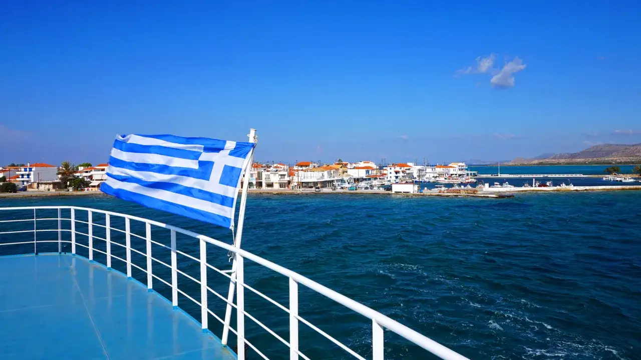 Griechische Flagge weht auf einem Schiff, das sich einem malerischen Küstenort nähert. Der Blick geht über das blaue Meer auf weiße Häuser und einen kleinen Hafen. Ein perfekter Start für eine Reise mit dem Zug nach Griechenland.