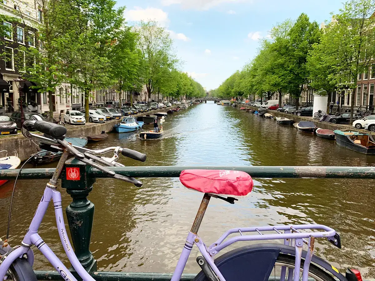 Ein lila Fahrrad mit rotem Sattel steht auf einer Brücke über einem Kanal in Amsterdam. Boote und Bäume säumen das Wasser, typische Amsterdam Attraktionen.