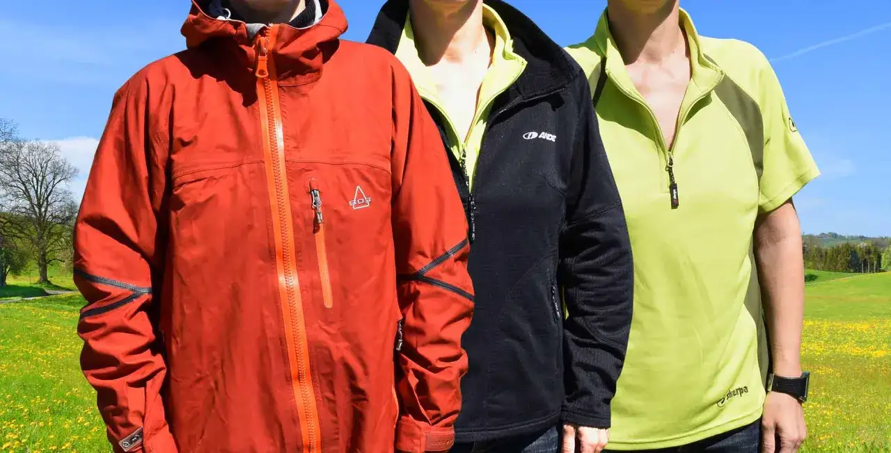 Drei Personen im Zwiebel-Look: rote Jacke, schwarzer Fleece und hellgrünes Shirt vor blauer Himmel und Blumenwiese.