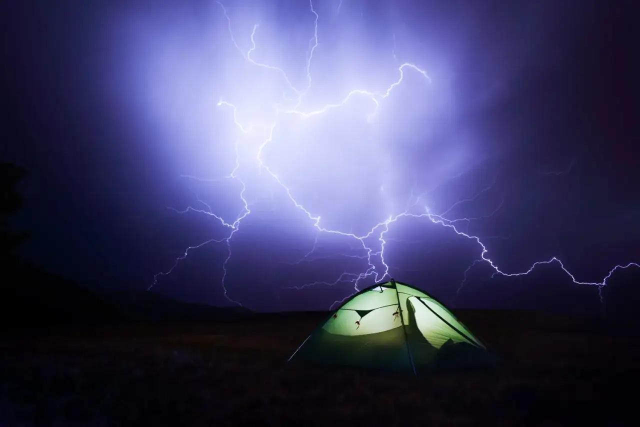 Ein gr&uuml;nes Zelt steht auf einer Wiese, w&auml;hrend ein dramatisches Gewitter mit Blitzen den Himmel erhellt.
