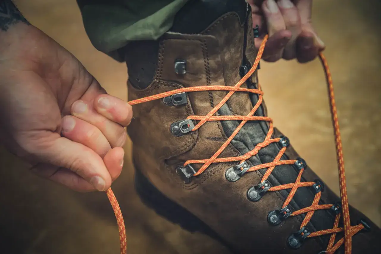H&auml;nde binden orange Schn&uuml;rsenkel an braunen Wanderschuhen. So bindet man Wanderschuhe richtig f&uuml;r den n&auml;chsten Ausflug.