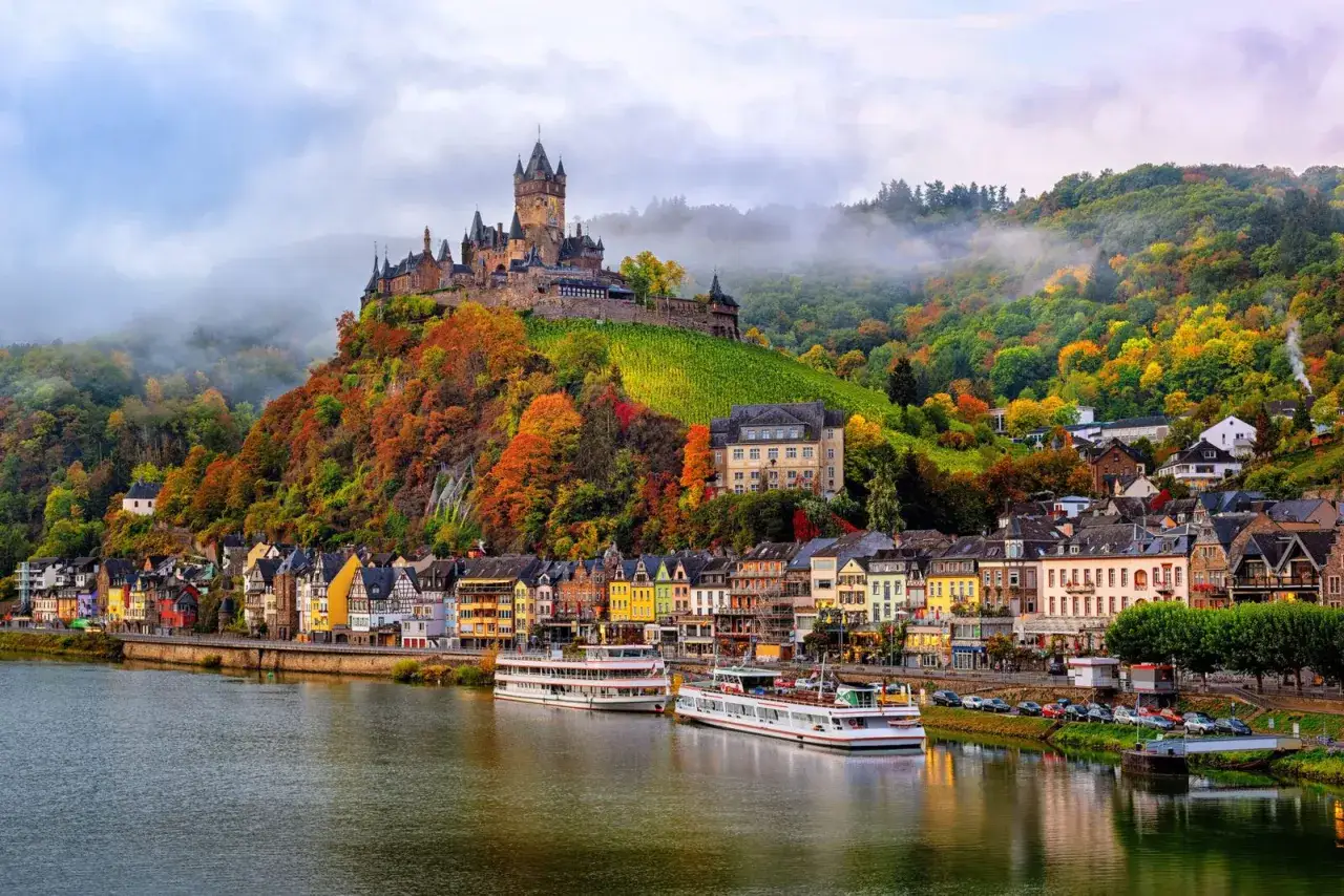 Malerische Burg thront &uuml;ber einem Dorf am Fluss, umgeben von bunten Herbstfarben. Ein Blick, der Fragen aufwirft, wie viele Burgen es wohl in Deutschland gibt.