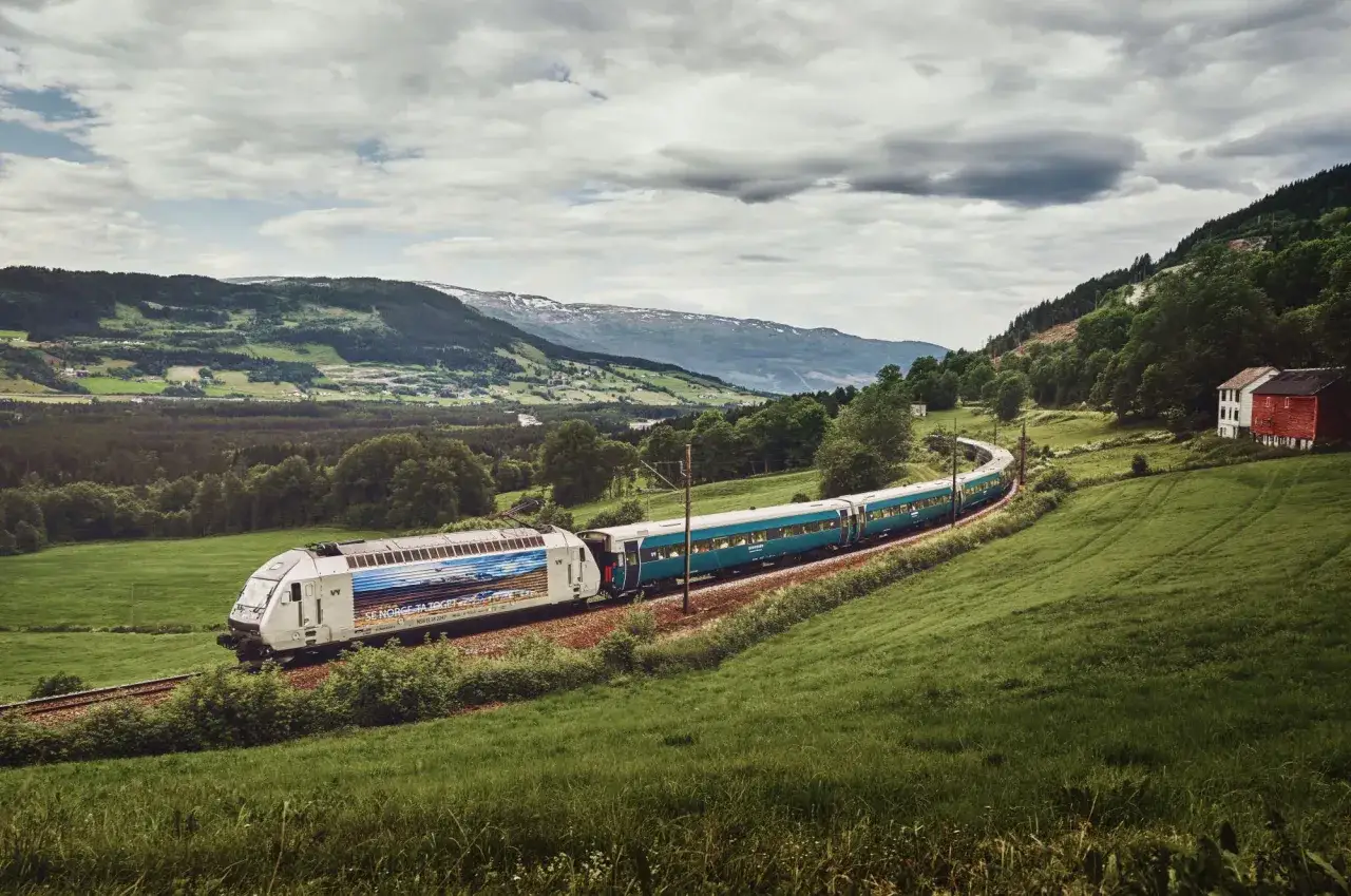 Die Bergen Bahn schl&auml;ngelt sich durch gr&uuml;ne H&uuml;gel und W&auml;lder, vorbei an roten H&auml;usern und schneebedeckten Bergen.