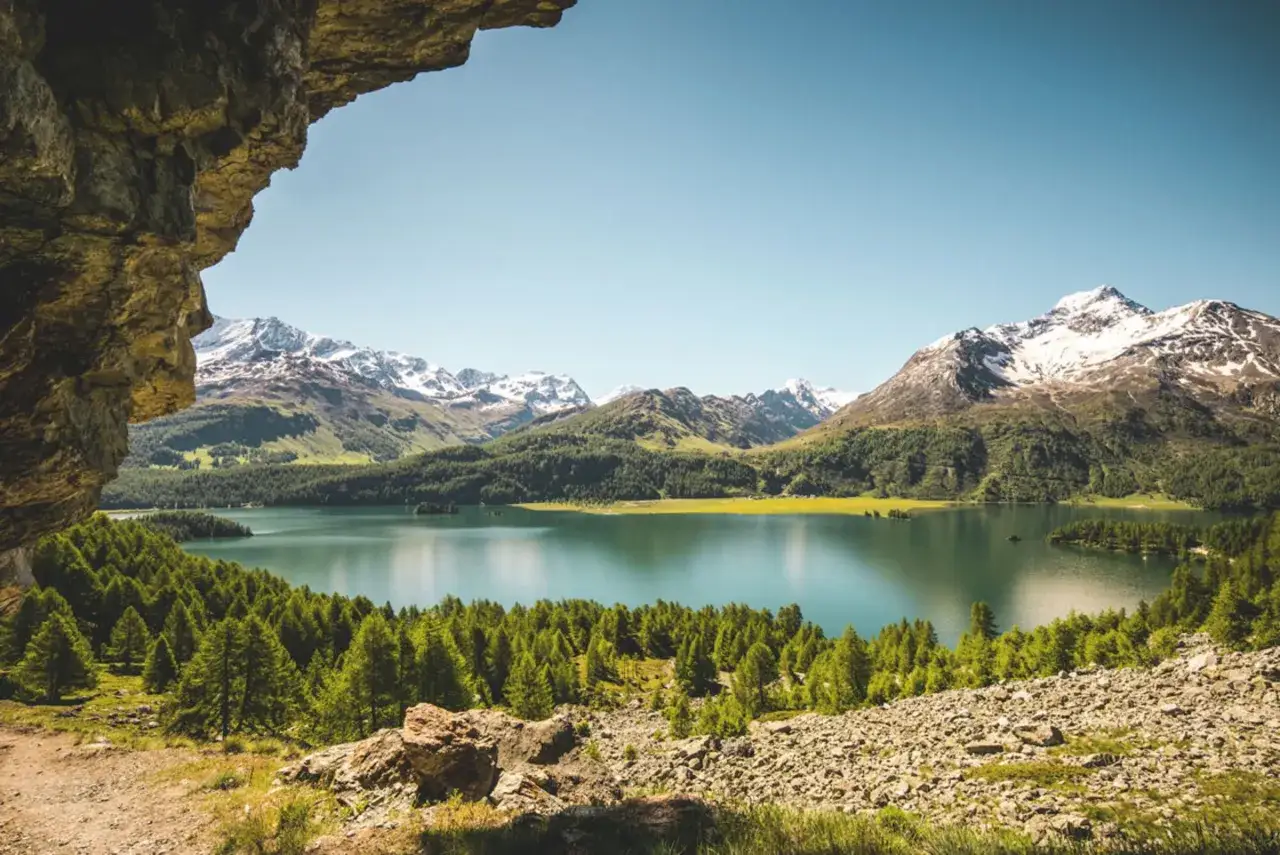 Felsiger Aussichtspunkt mit Blick auf einen t&uuml;rkisfarbenen See, umgeben von gr&uuml;nen W&auml;ldern und schneebedeckten Bergen. Ein malerischer Ausblick, der an die Grenze zwischen Schweiz und Italien erinnert.