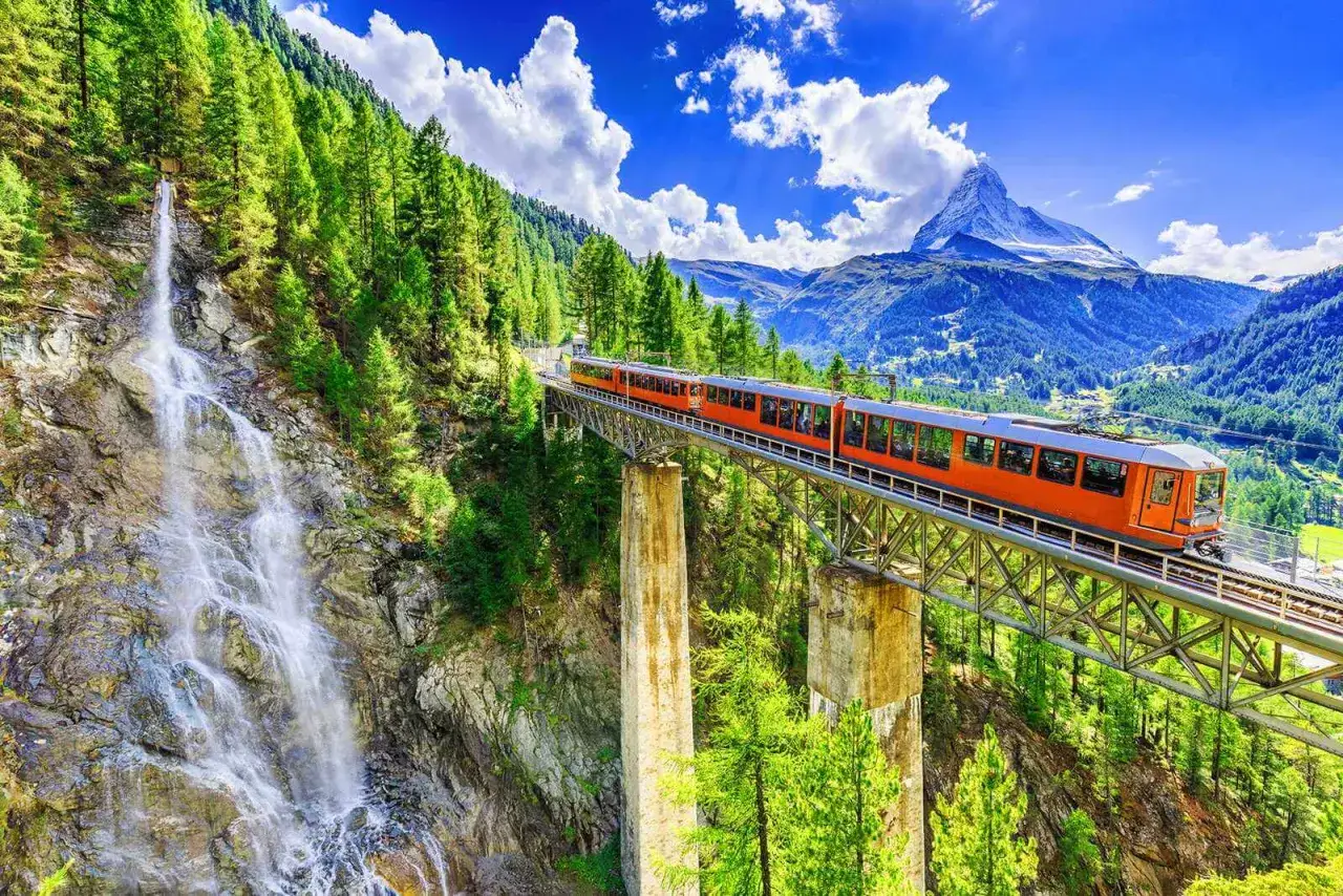 Orange train crosses a viaduct near a waterfall in the Alps. A perfect scene for exploring the beauty of festivals Europa.