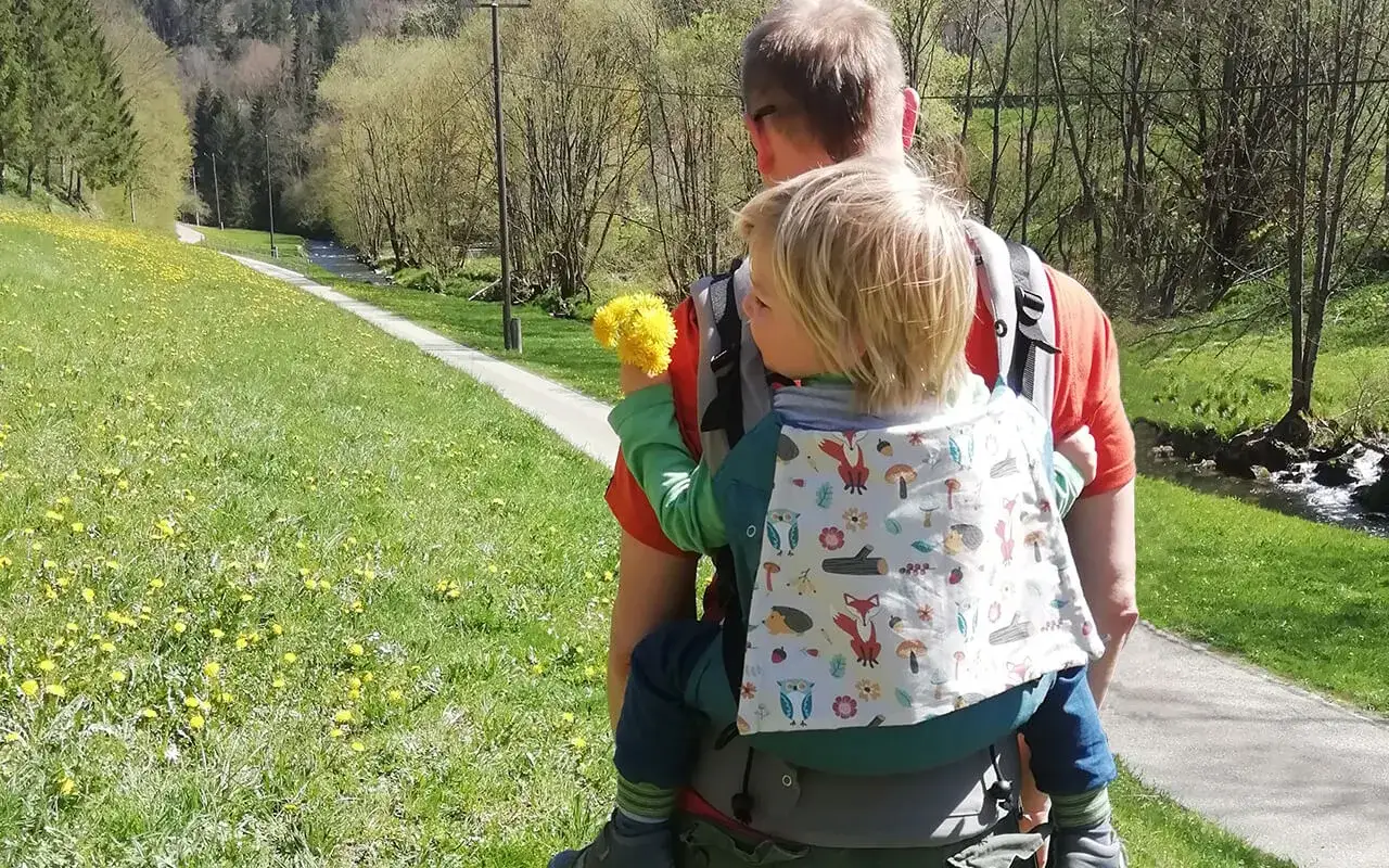 Ein Vater genie&szlig;t das Wandern mit Baby auf einem sonnigen Weg entlang eines Baches. Das Kind h&auml;lt eine L&ouml;wenzahn-Wolke.