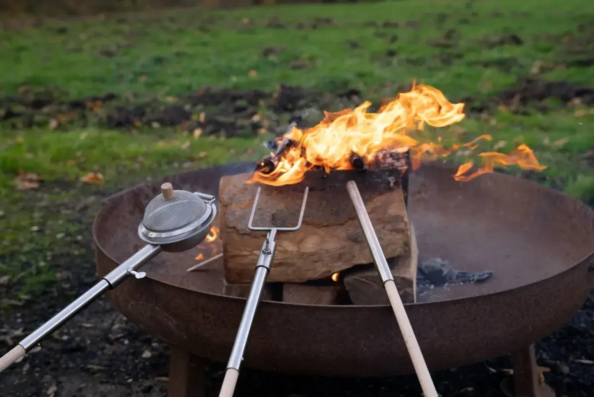 Ein gem&uuml;tliches Lagerfeuer machen: Holz brennt in einer Feuerschale, daneben liegen ein R&ouml;stsieb und eine Grillgabel.