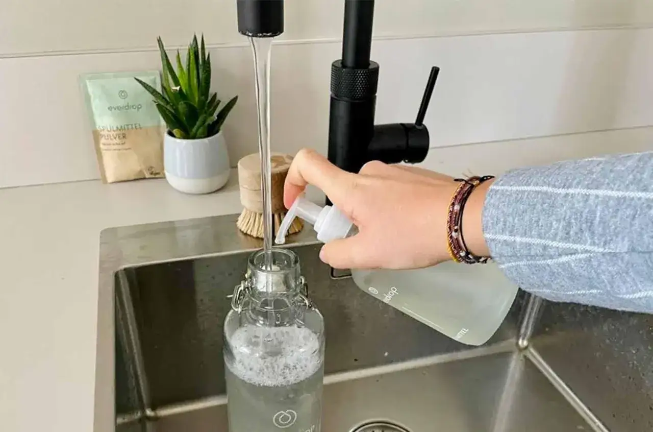Wasser fließt in eine Glasflasche, während eine Hand Spülmittel hinzufügt. So einfach lässt sich die Edelstahlflasche reinigen.