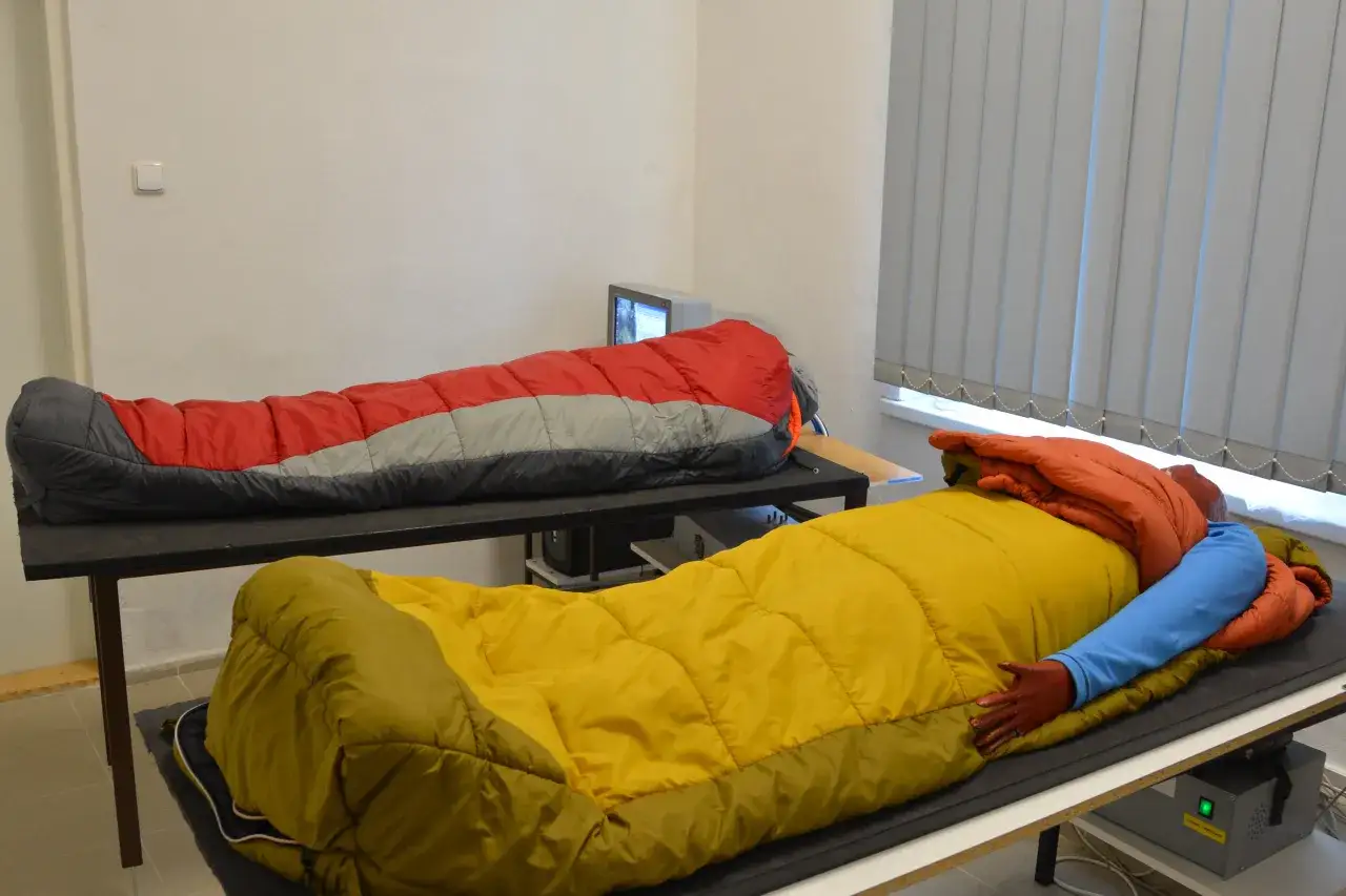 Zwei Schlafsäcke liegen auf Tischen. Einer ist rot-grau, der andere gelb. Ein Arm ragt aus dem gelben Schlafsack, um die schlafsack komforttemperatur zu testen.