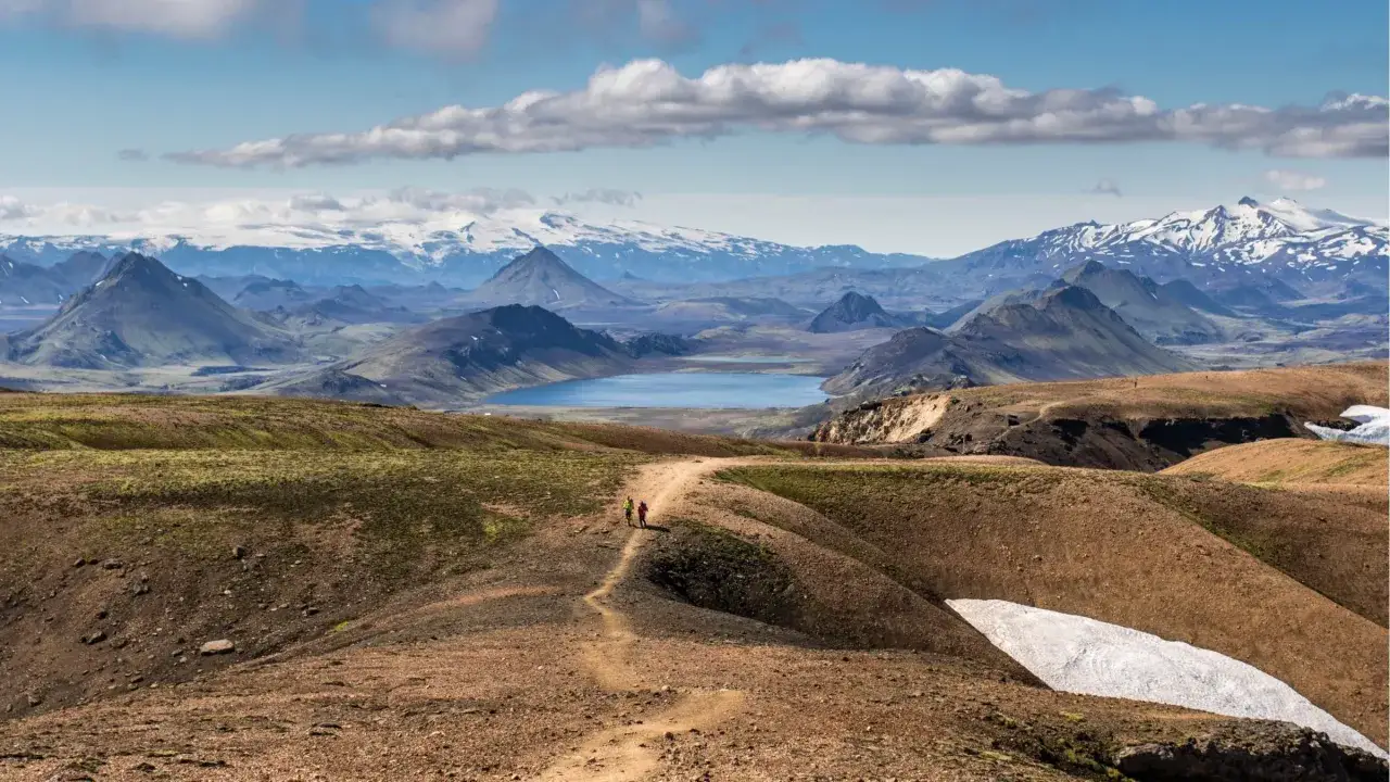 Zwei Wanderer auf einem Pfad in einer bergigen Landschaft mit einem See und schneebedeckten Gipfeln im Hintergrund. Perfekt für mehrtageswanderungen Europa.