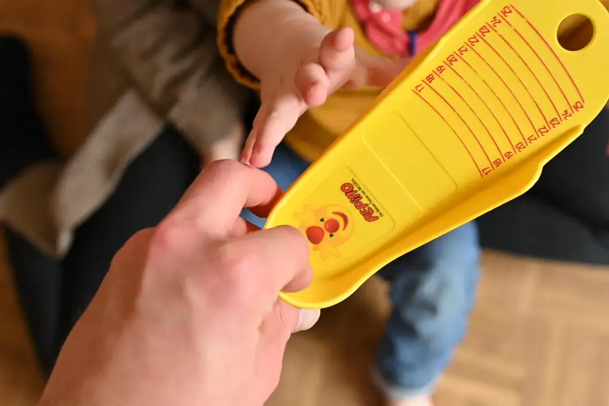 Ein gelber Fu&szlig;messer zeigt, wie fallen Trollkids Schuhe aus. Eine Hand h&auml;lt ihn, ein Babyfu&szlig; wird gemessen.