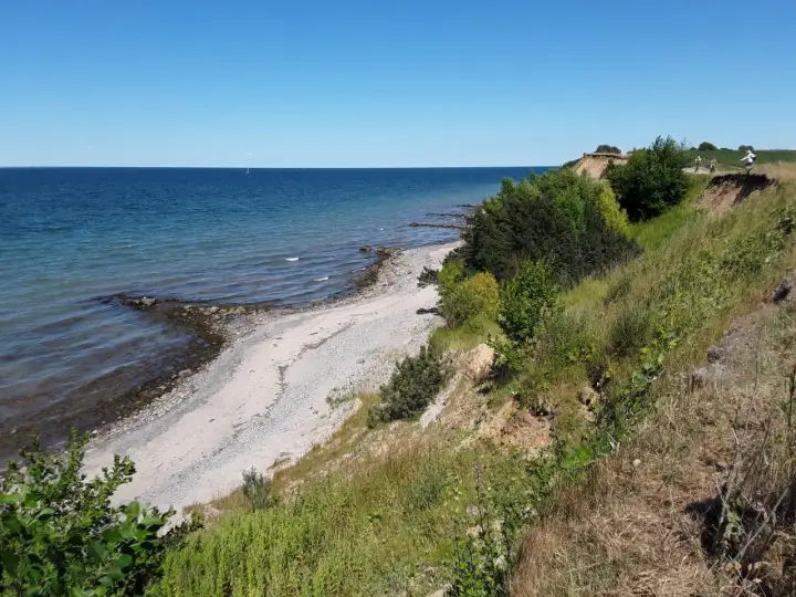 Ein sonniger Tag am Timmendorfer Strand. Die Ostsee glitzert, ein schmaler Kiesstrand lädt zum Spaziergang ein, während grüne Büsche die Steilküste säumen.